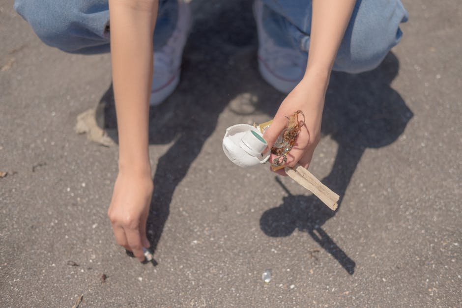 A person wearing light blue jeans and white sneakers is crouched on a paved surface, holding a small pile of mixed rubbish including a crumpled white paper cup, a wooden stick, and some tangled string