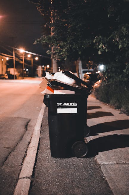 A black wheelie bin positioned on a sidewalk at night, labeled with the name 'St. John's' and a blank space for additional identification. The bin contains various types of waste including flattened c